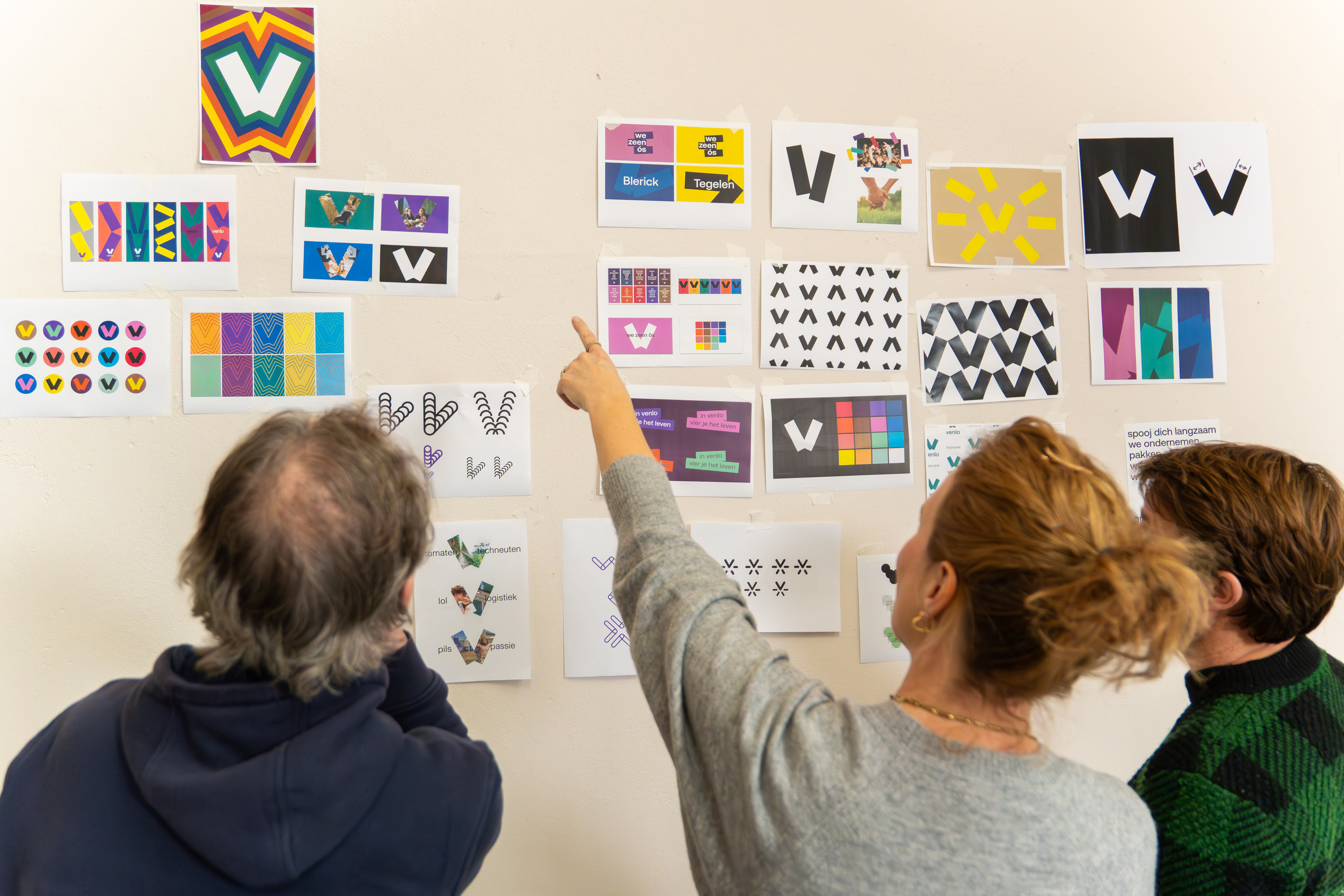Three people stand looking at a wall with different examples of Venlo's place brand identity stuck to it. The woman in the middle is raising her finger to direct attention to a specific design example. The Venlo design is a blocky ''V' made up of two rectangles, with a bright colour palette. 