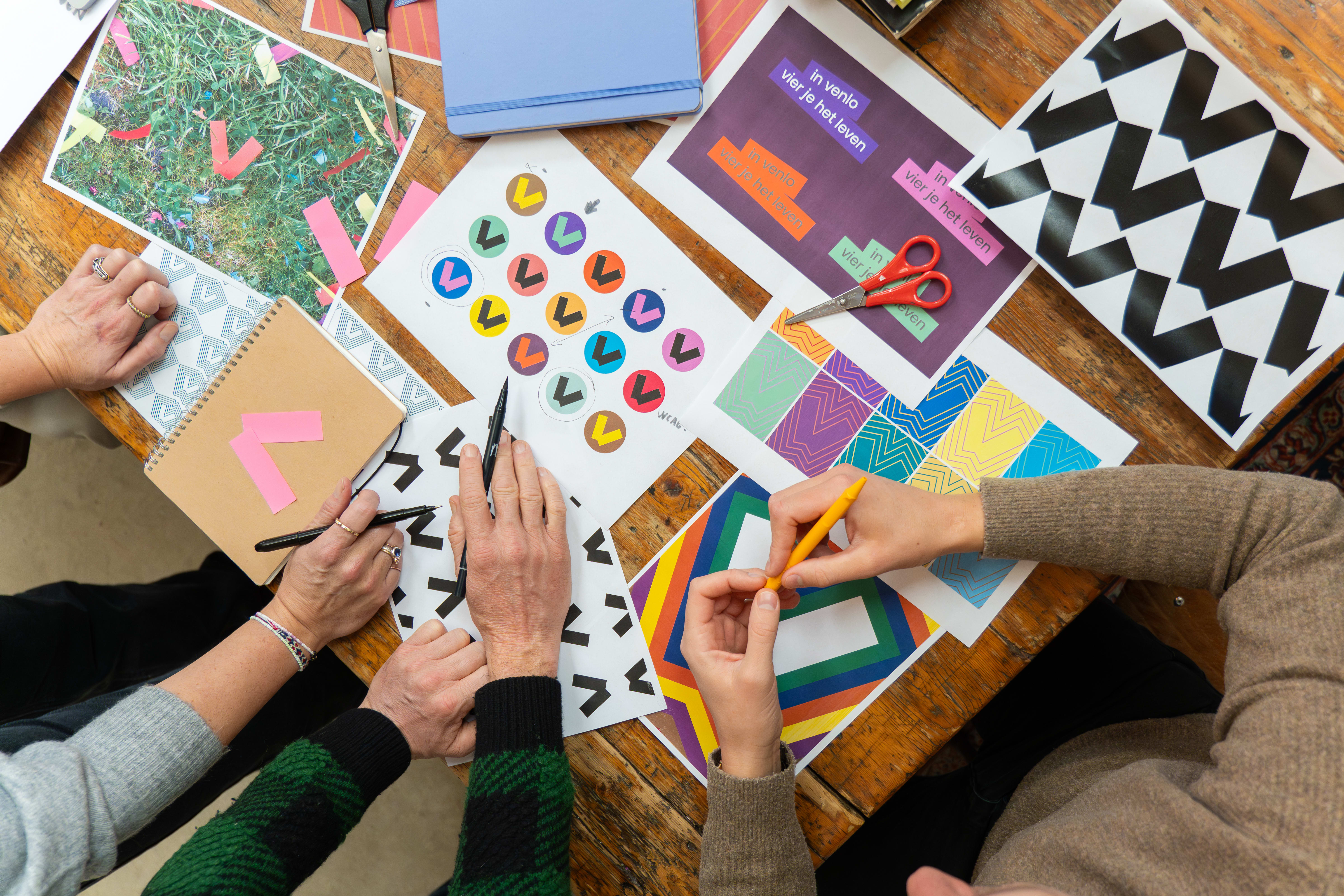 An overhead shot of a table covered in examples of Venlo's branding. Venlo's branding is a 'V' made up of two blocky rectangles, and the examples show how the logo can be used as an icon, a background, and more. The arms of three people can be seen in the bottom of the shot as they discuss the designs.